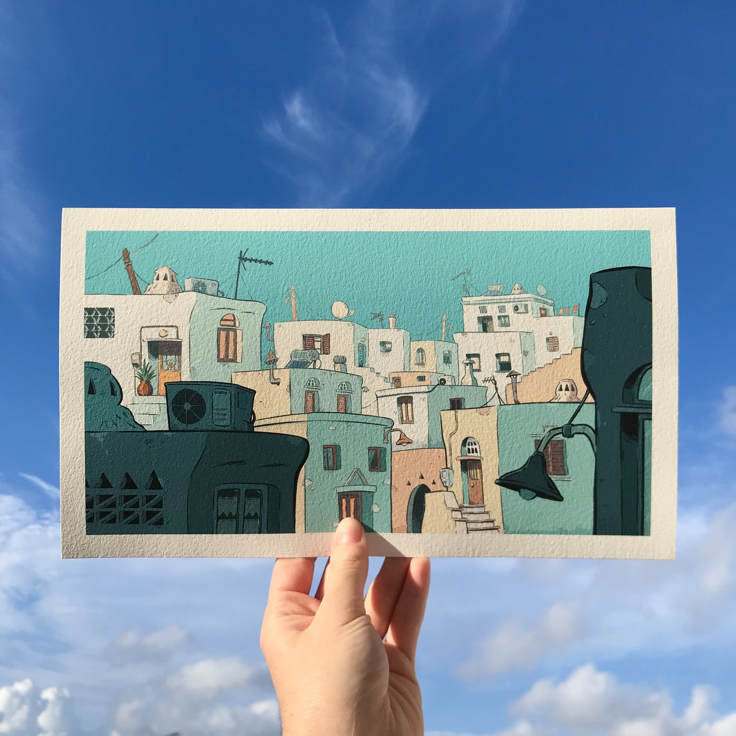 Illustration hold by a hand with the sky in the back. The theme of the work is a cycladic village in shades of blue and yellow and beige.