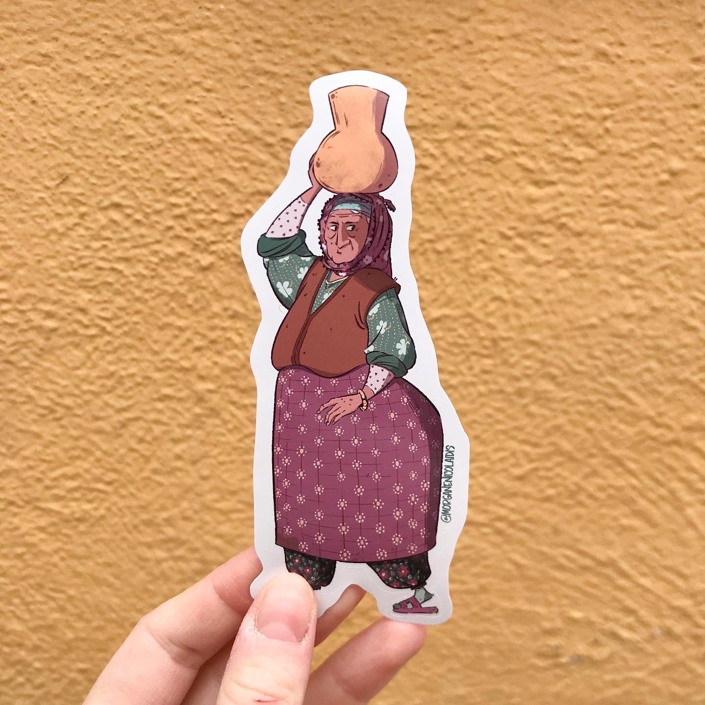 The sticker is hold with a hand in front of a yellow wall.