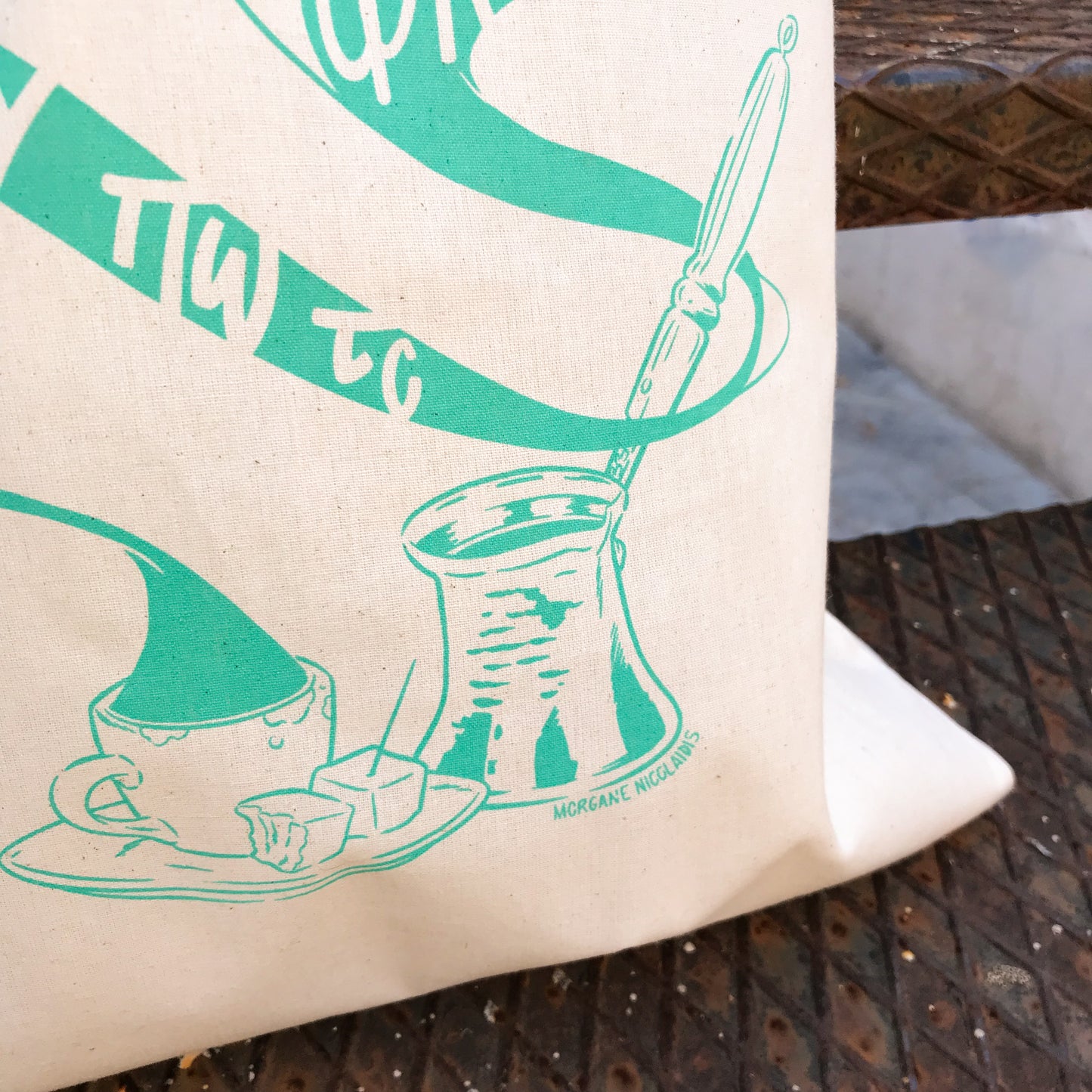 Close up of the coffee and the briki. The tote bag is on some metallic stairs.