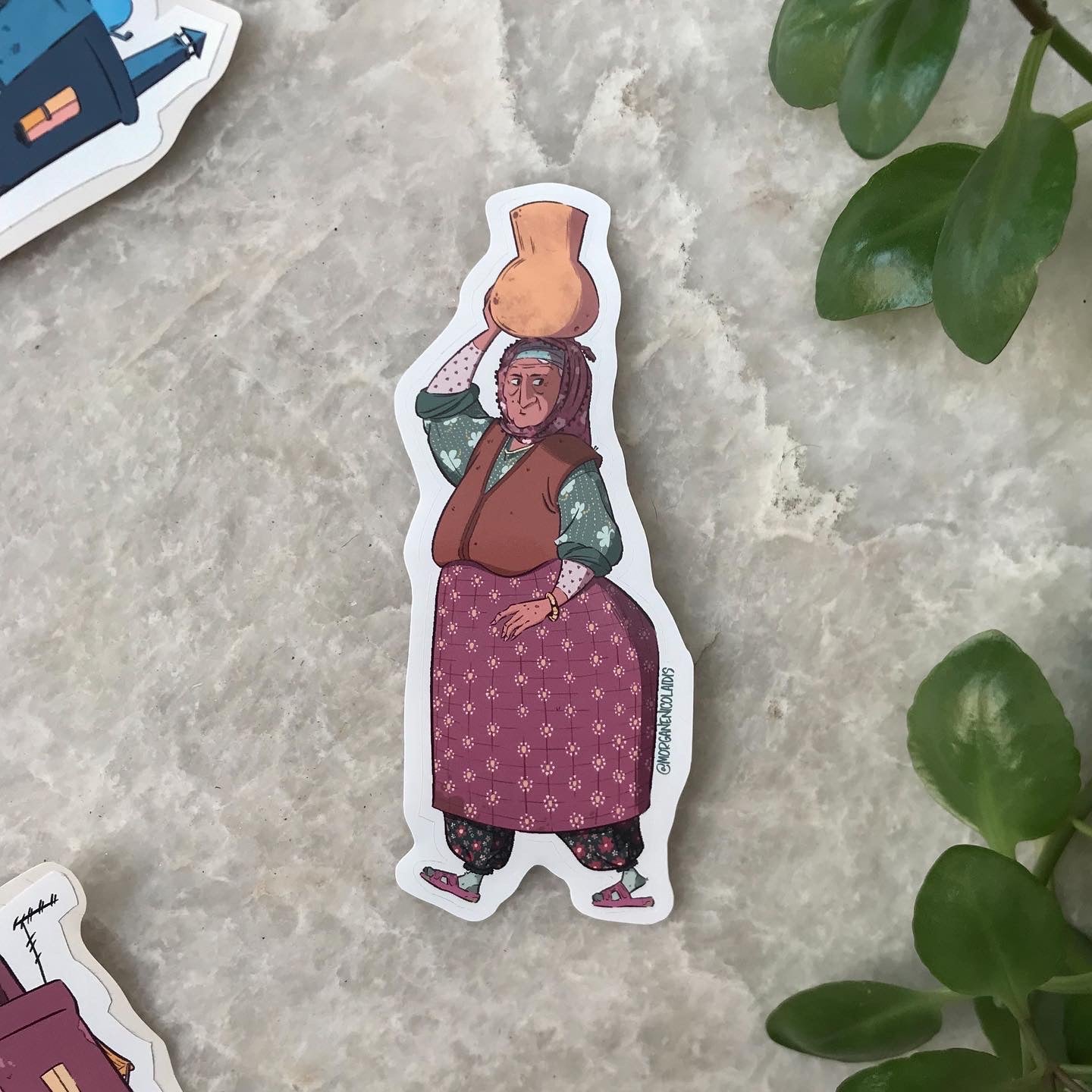 The sticker is placed on a marble floor, surrounded by stickers and a few leaves.