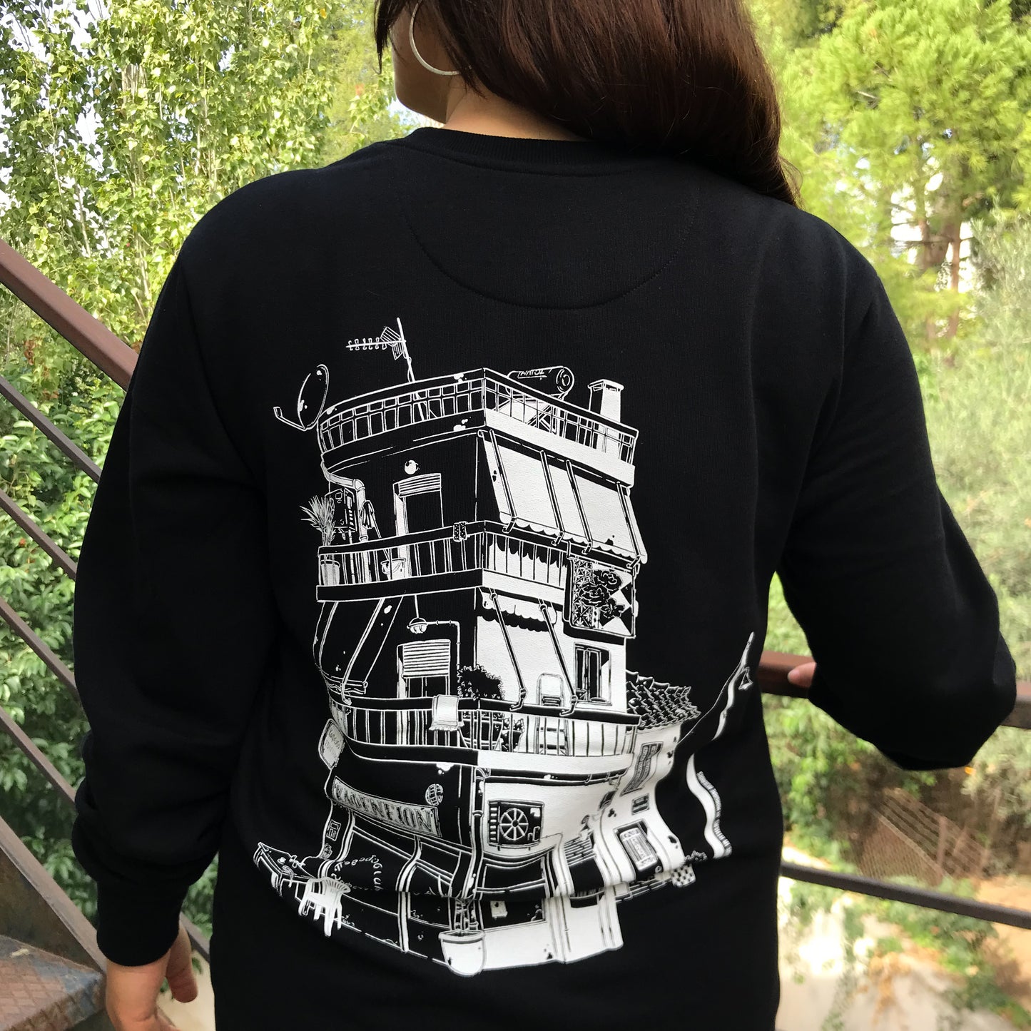 close up of this black sweatshirt depicting a Athenian/ Greek building only with a white lineart. On the building we have different details such as yucca plant, a "Kafeneio" on the ground floor, big blanket hanging on a balcony, a smaller old house at the right of the building., and satelite antenna.