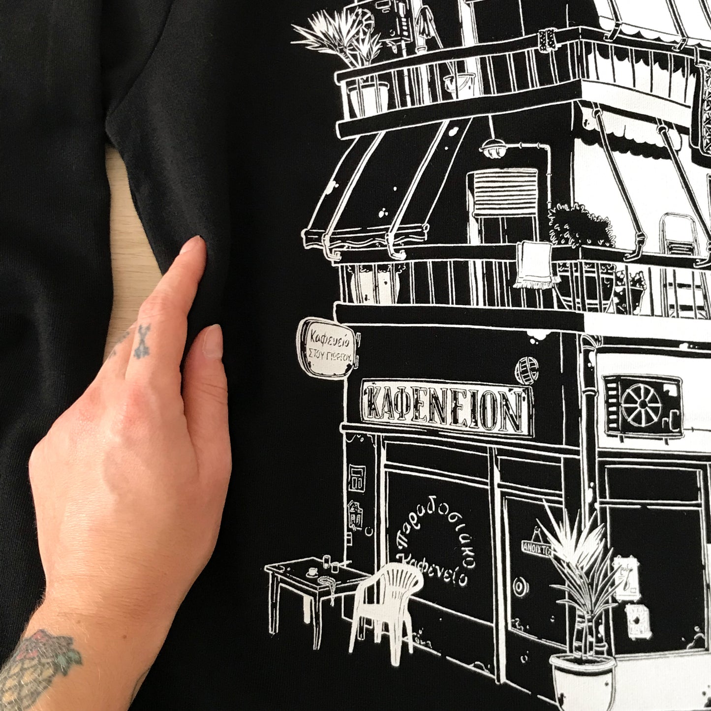 close up of the back design (left part) with a hand touching the fabric. The details visible are the "Kafeneio" with a plastic chair and a table, some yucca plans, balconies with towel hangings, an air conditioning and some inscription in greek (such as "traditional kafeneio").