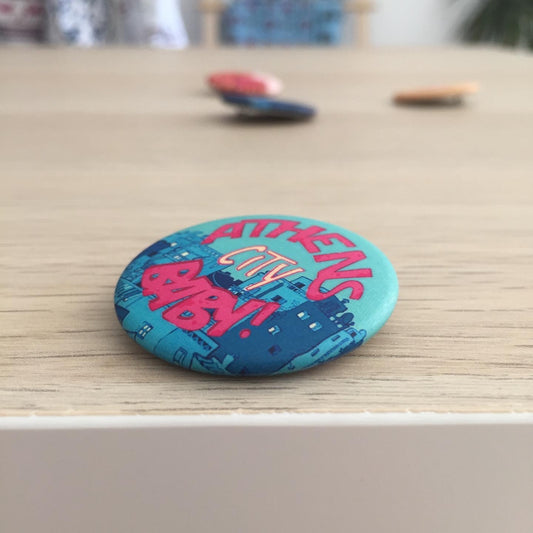 Button pin on wooden table. Athens City Baby is written, and a city scape is drawn in the background