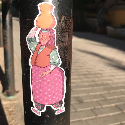 This is a sticker of a woman from Anatolia (Anadolu, in Turkey). She is wearing colorful clothes, a headscarf, and shes's holding a jar of water. The sticker is sticked on metallic pole in the street.
