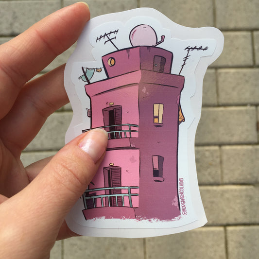sticker of a pink building inspired by greek architecture hold by a hand. The background is a cobbled street.