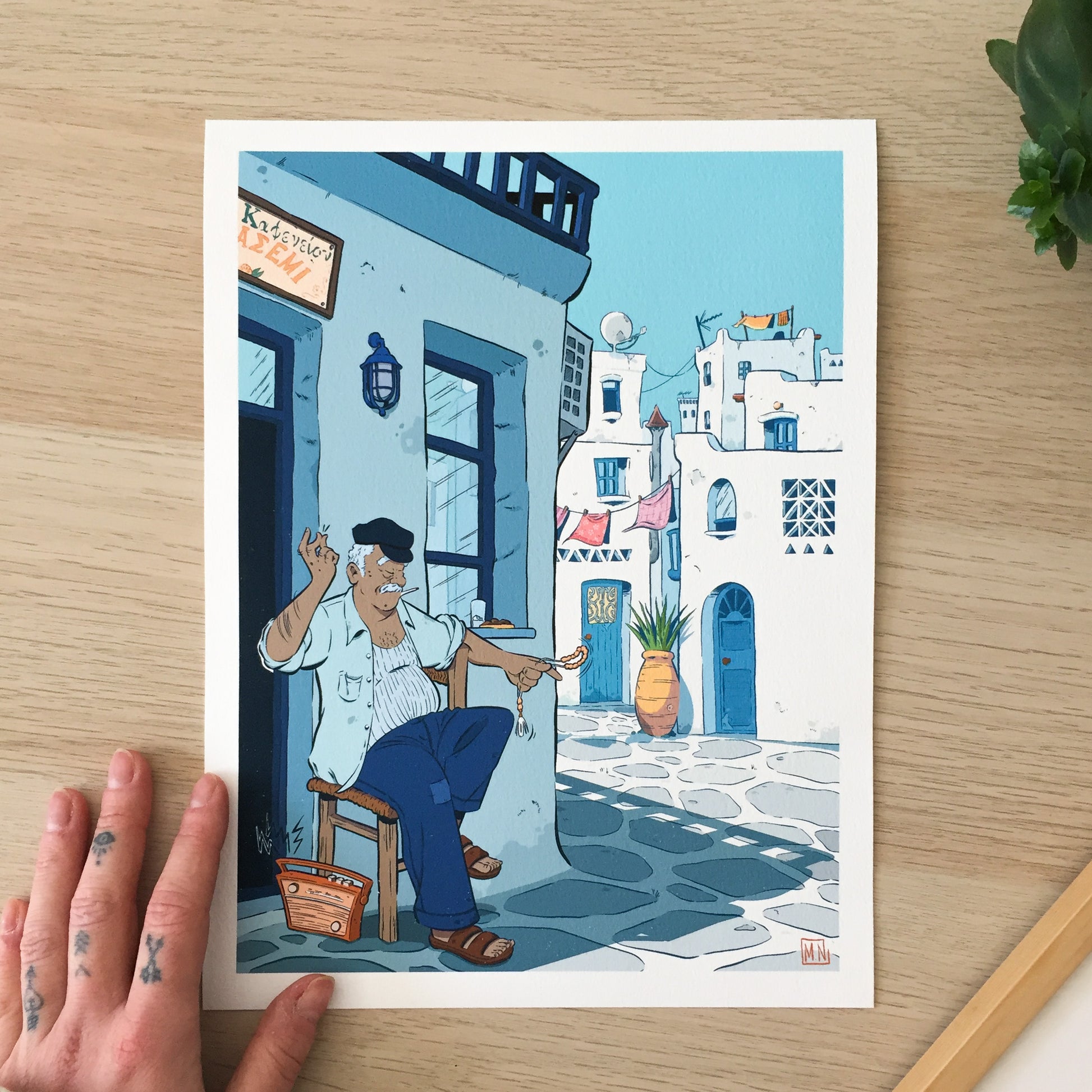 A Kafeneio, in blue colours, with a man at the left down corner of the image. He is listening to some music from a radio display while clapping his finger. He has a komboloï on his left hand. The village in the background is a typical cycladic village.
