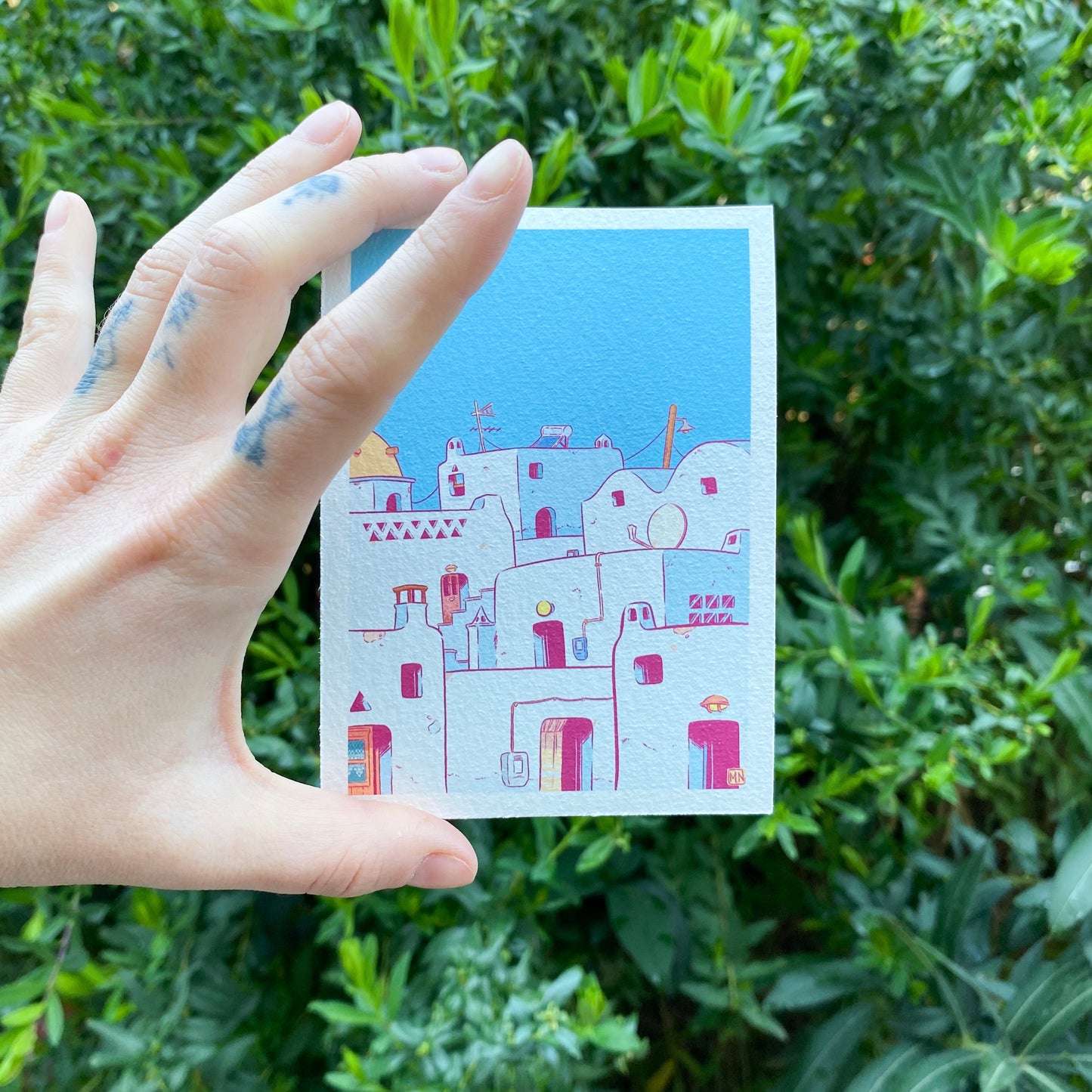 The mini print is hold by a hand in front of some bushes.