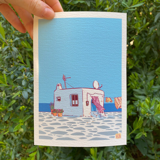 a small print hold by a hand with some plants as the background. The print is the illustration of a cycladic house in front of the sea. We have some laundry hanging on a a wire and a curtain. An exterior curtain hung on the front door. A few plants are displayed near the house.