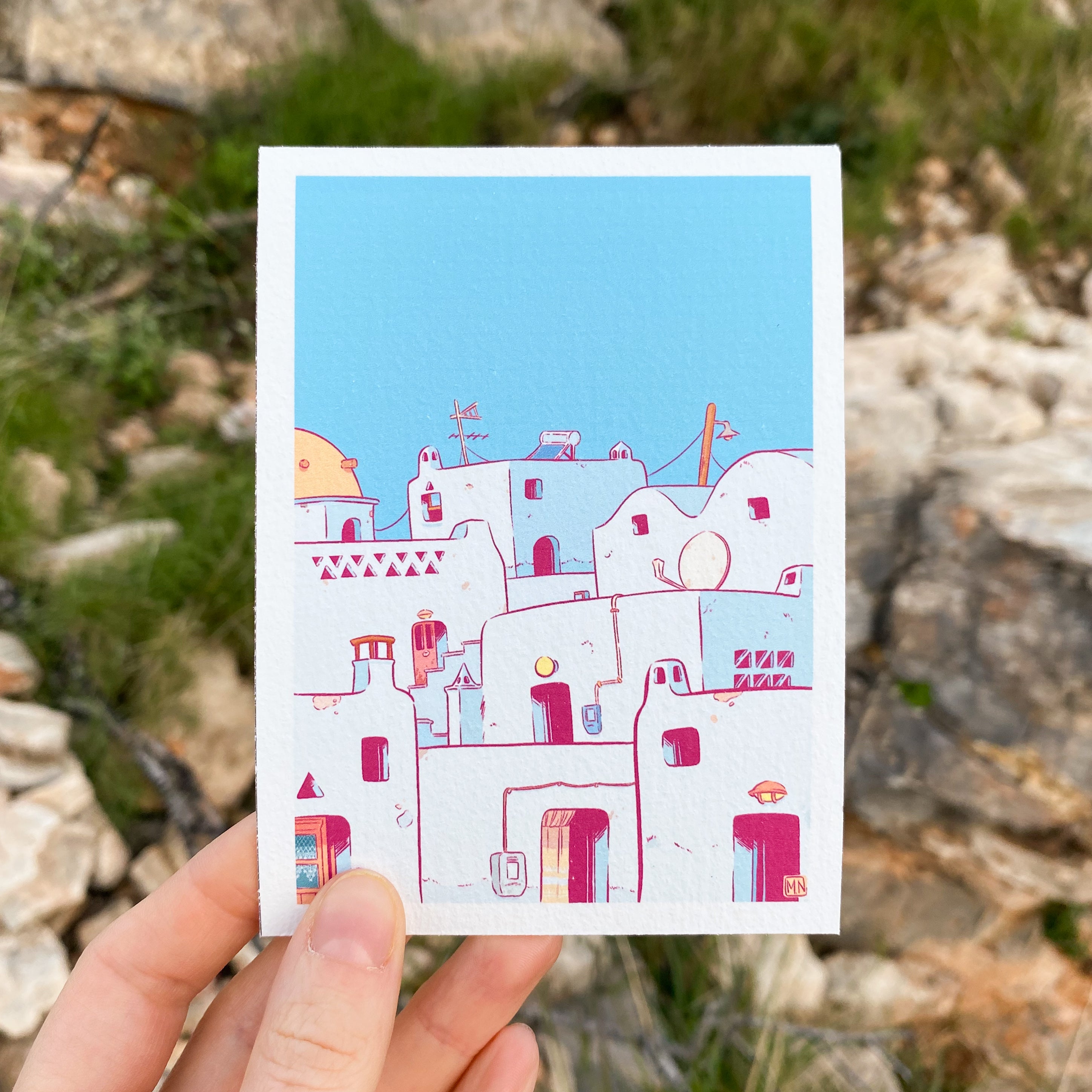 A mini print of a greek cycladic village hold by a hand. At the back of the print we have some rocks and grass.