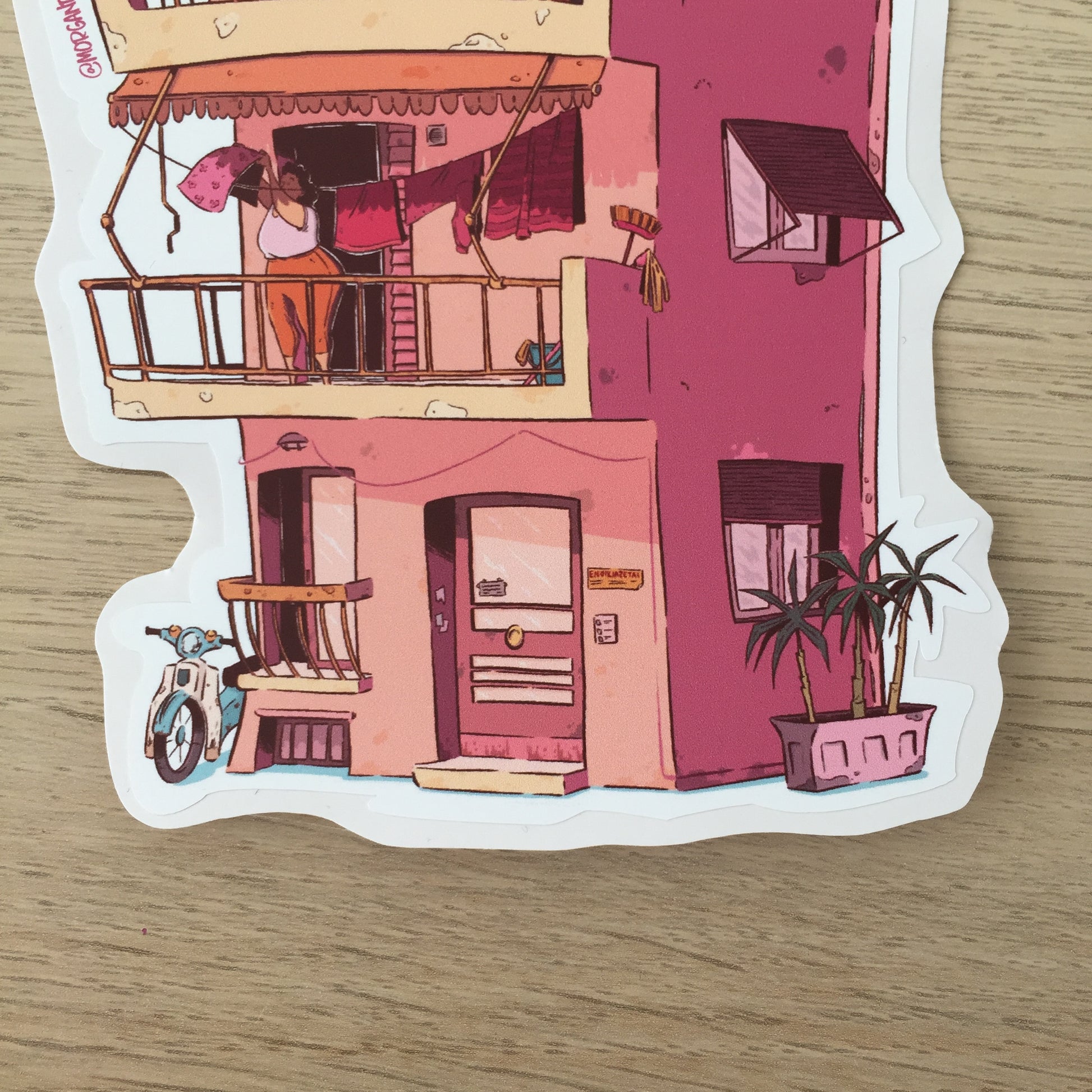 Close up of the bottom of the sticker, where we can see a lady hanging out her laundry, there is a motorcycle at the left bottom corner, there is also three little plam trees at the right of the building.