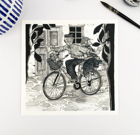 Old couple in love on a bicycle. The background is a greek village road, with an old door of a traditional house. We have two trees on the first plan of the image. The illustration is on a white table with a brush and indian ink container on the top right pof the photo and a a flower pot on the top left.