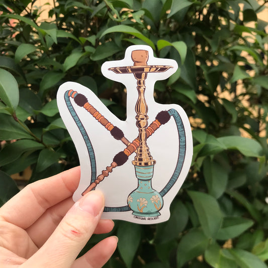 A hookah pipe, in blue, turquoise and gold, hold by a hand with a bush as a background.